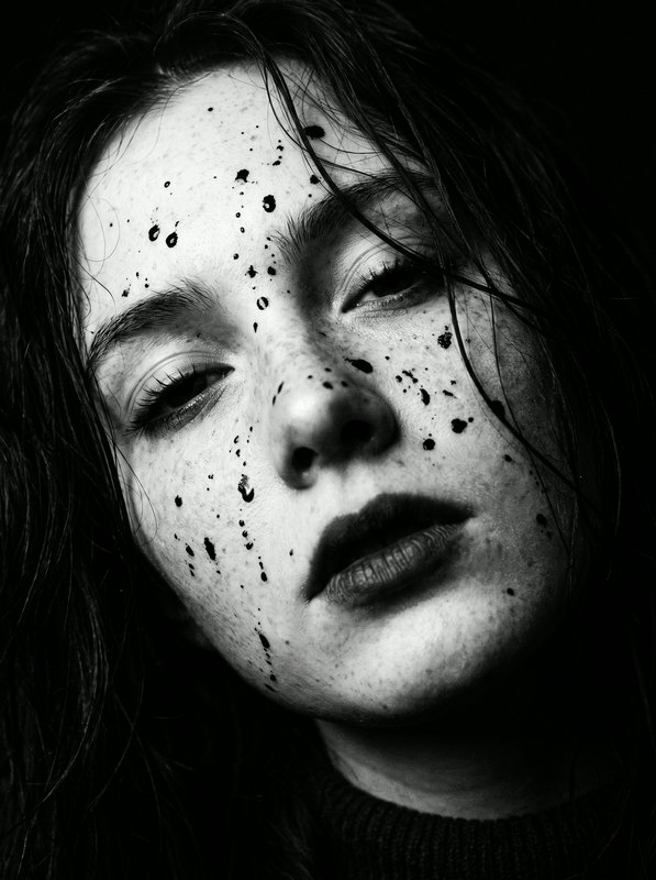 Close-up monochrome portrait of a young woman with dark paint splatters trailing down her face, m...