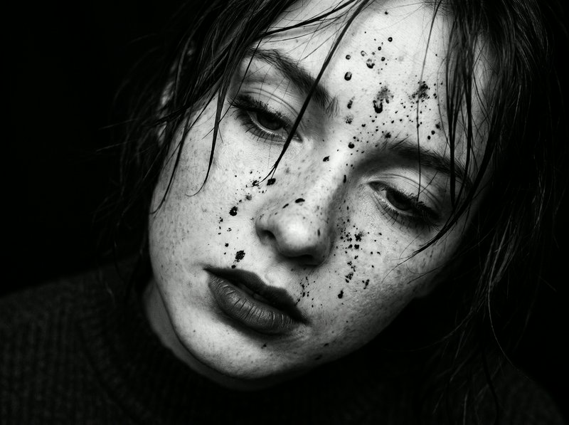 Close-up monochrome portrait of a woman with wet hair, dark splatters across her face, and a somb...