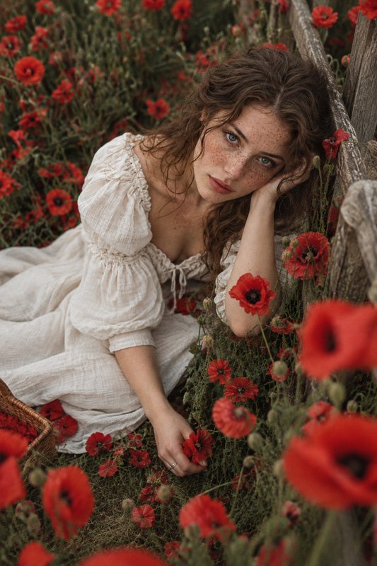 Freckled woman in a cream prairie dress resting against a wooden fence in a field of red poppies