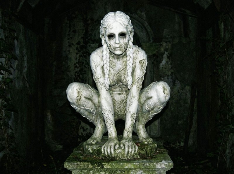 Creepy crouched stone female statue with hollow eyes and braided hair rests on a mossy pedestal in a dark ruined crypt