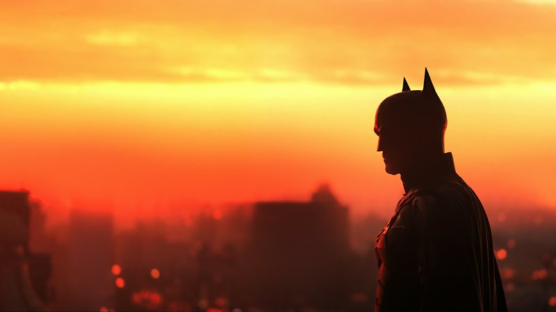 [Side profile silhouette of Batman against a vivid orange sunset over a blurred Gotham City skyline]