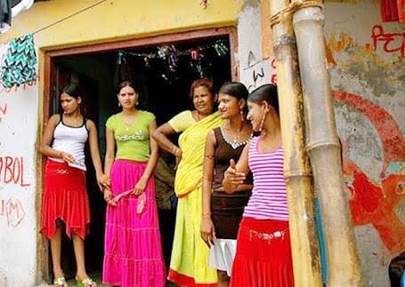 Five South Asian women stand casually together in the doorway of a colorful painted residential neighborhood home.
