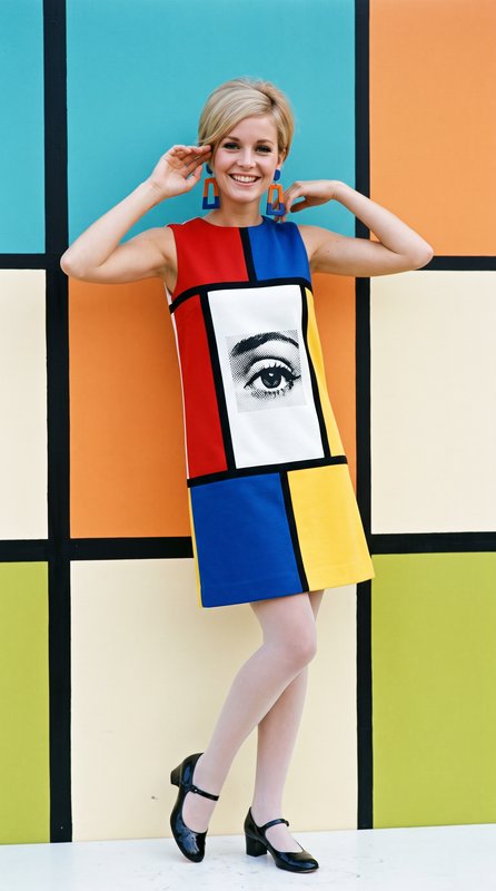 Cheerful 1960s mod model wears a Mondrian grid shift dress with pop art eye graphic, posing against matching color block w...