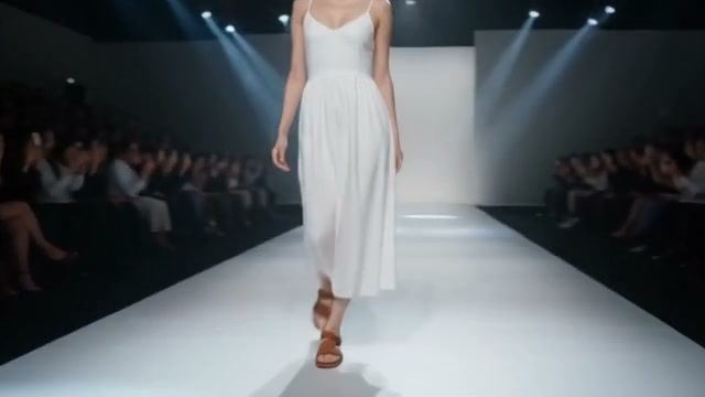 Model in a flowing white spaghetti strap midi dress walks down a lit fashion show runway as the f...