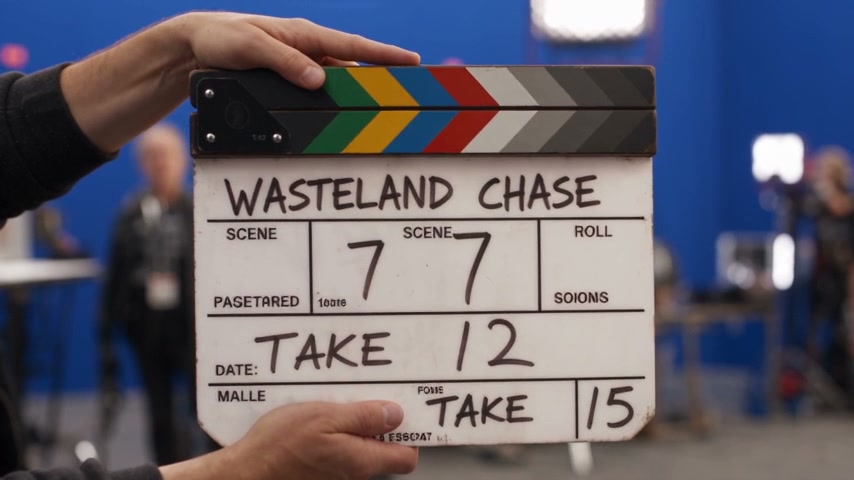 Hands holding a colorful striped film clapperboard marked "Wasteland Chase" on a blue screen movi...