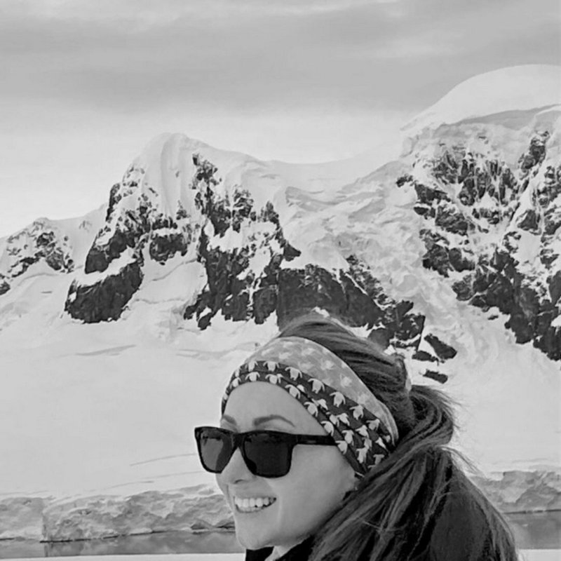 Smiling woman in sunglasses and patterned headband stands before snow-covered glacial mountains