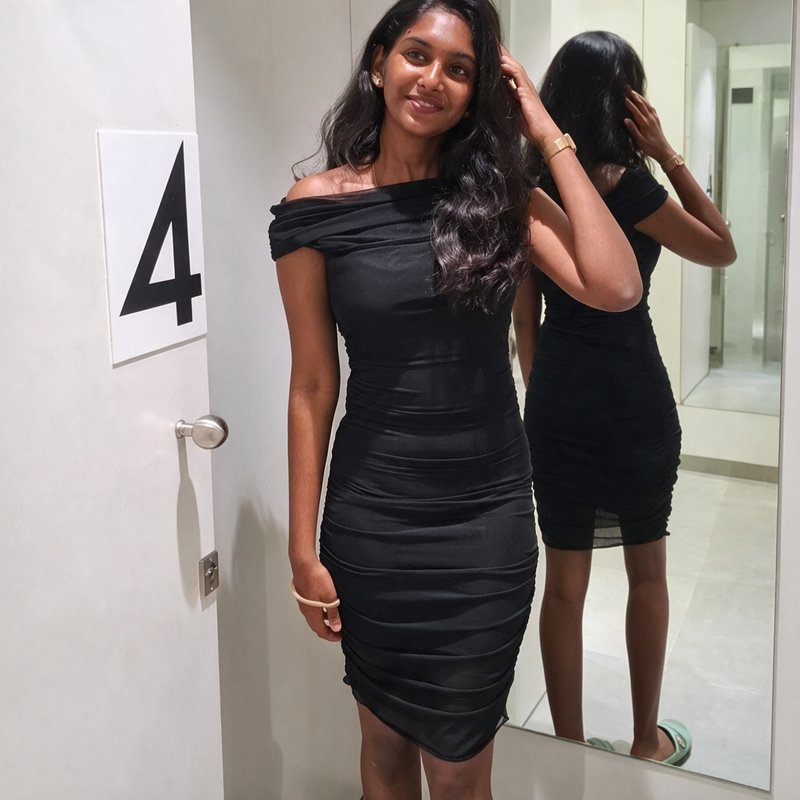 Smiling woman trying on a ruched black off-shoulder dress in fitting room number 4
