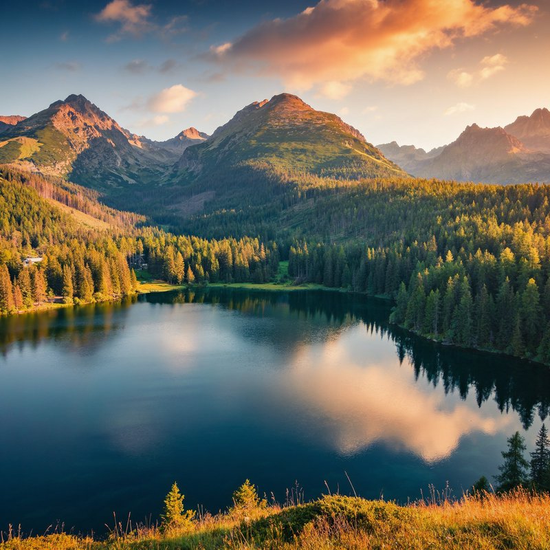 Calm alpine lake reflects forested mountain peaks and golden hour clouds in a peaceful mountain v...