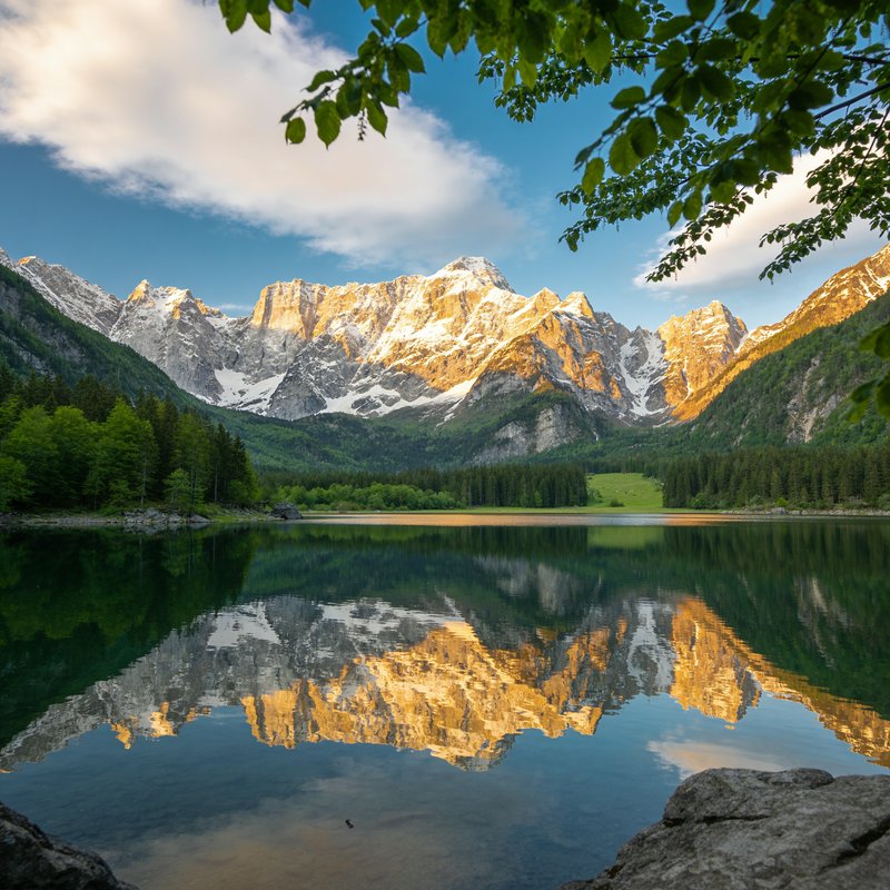 Glassy alpine lake reflecting sunlit snow-capped peaks, framed by green tree foliage at golden ho...