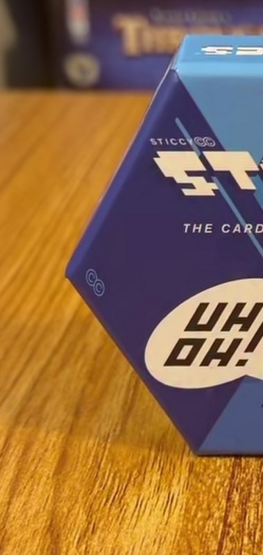 Close-up of hexagonal STICCY card game box sitting on warm grained wooden table with a soft blurr...