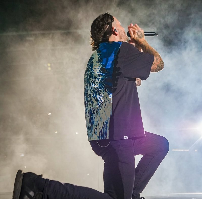 Tattooed male vocalist kneels singing into a microphone on a smoky lit concert stage during a liv...