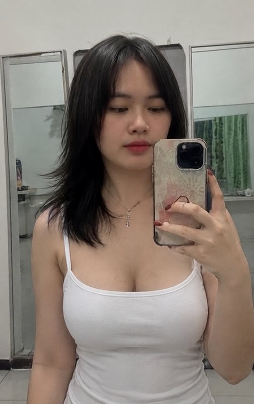 Young woman with layered black hair taking a mirror selfie wearing a white spaghetti strap top