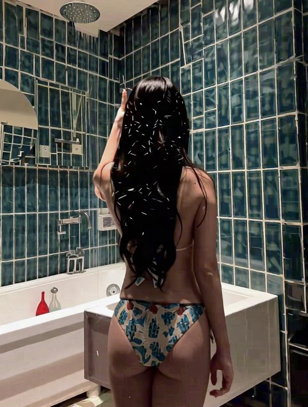 Rear view of woman in printed bikini standing under running overhead shower in teal tiled bathroom.