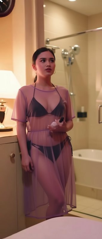 Young woman in black bikini and sheer lilac mesh coverup stands in warm lit luxury bathroom holdi...