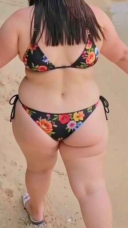 Rear view of a plus size woman in a black floral bikini standing on a sandy beach shore