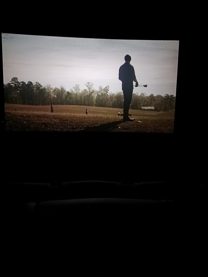 Silhouetted man holding a rifle stands in a sunlit rural field viewed on a home television screen
