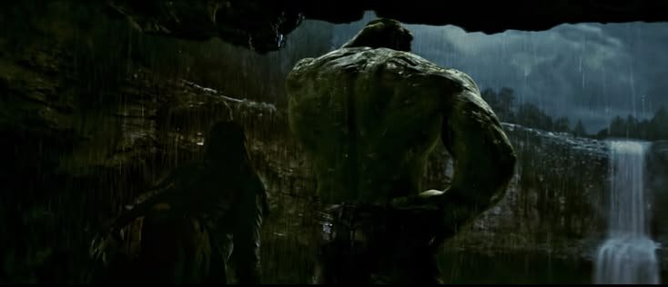Rear view of the muscular green Hulk standing under a rocky overhang during a rainy night, overlo...