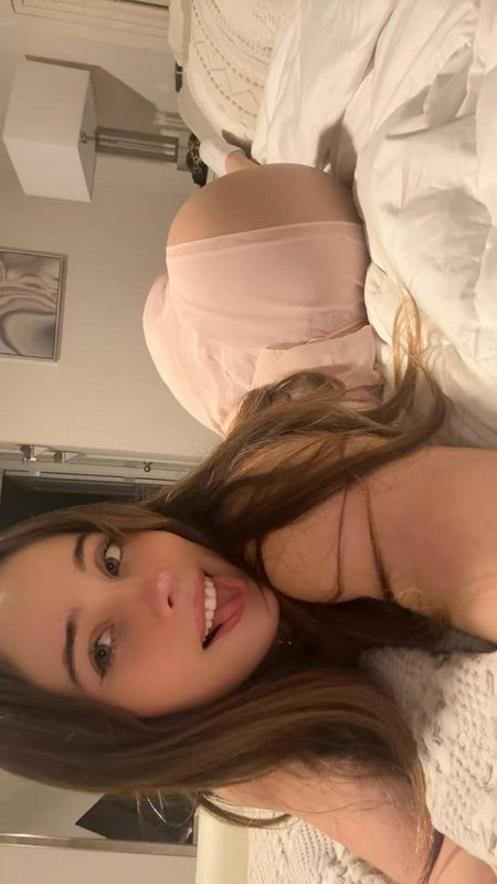 Playful young woman sticks her tongue out while taking a casual selfie reclining on a bedroom bed