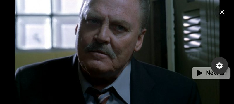 Close-up of a serious mustached older man in a suit staring intently at the camera in a dim office
