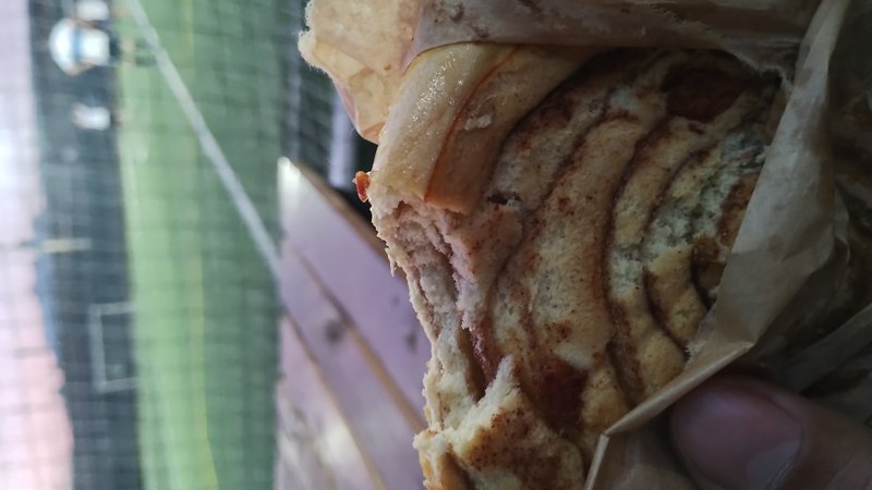 Partially eaten swirled cinnamon roll held in paper wrapping, outdoors near a sports field