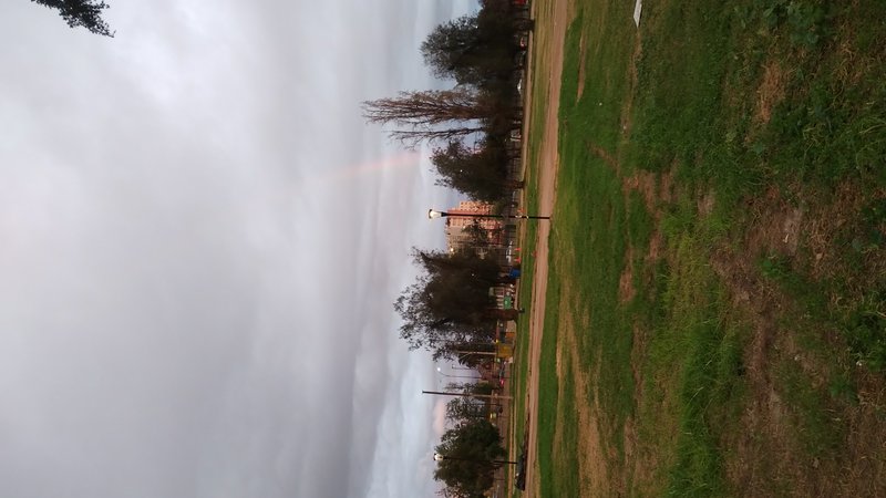 Faint pale rainbow arcs across a cloudy overcast sky above a grassy urban park at dusk