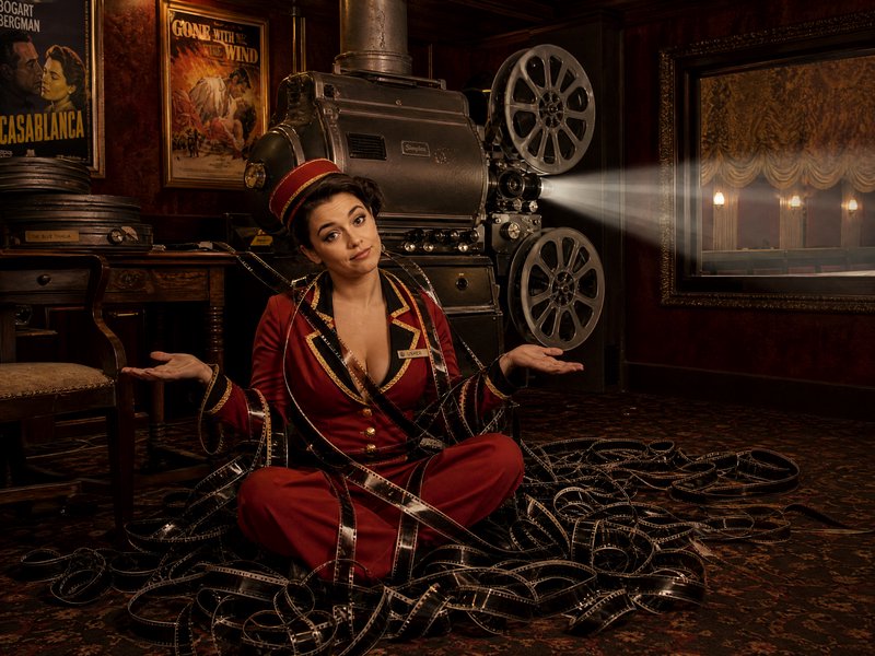 Woman in vintage theater usher uniform shrugs, tangled in 35mm film strips before an antique runn...