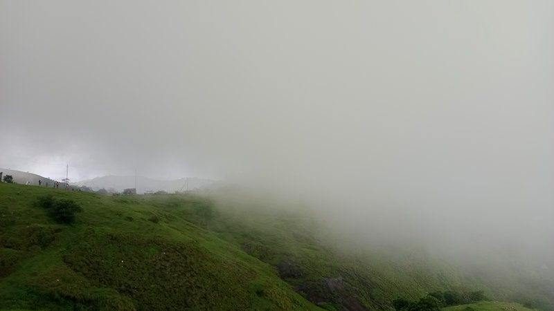 Thick fog drifts across a lush green sloped hillside with people on the distant ridge