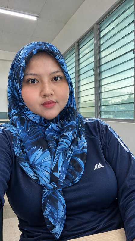 Selfie of a Muslim woman in a blue leaf-patterned hijab and navy athletic top, seated indoors nea...