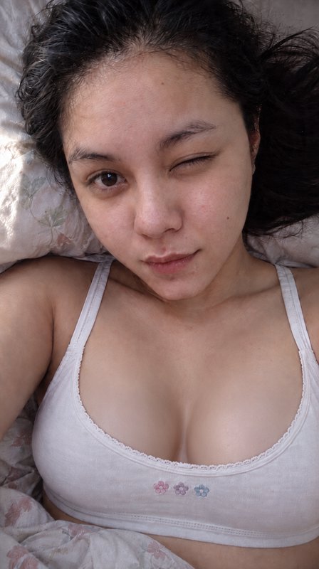 Young woman lying in bed winking playfully while taking a casual morning selfie in a soft embroid...