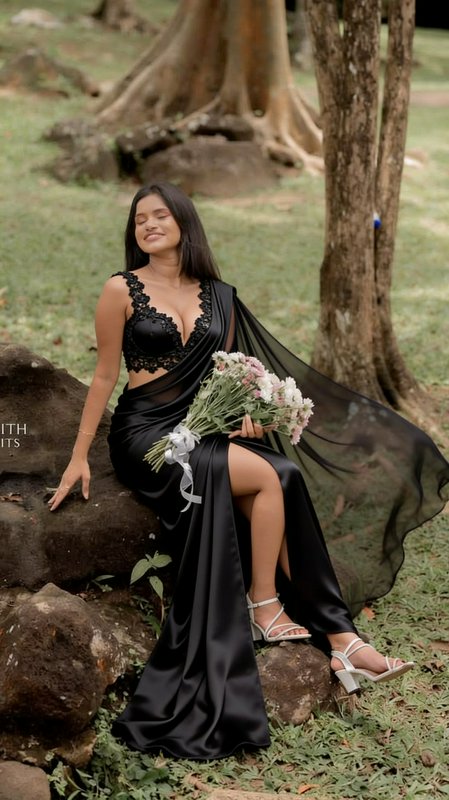 Smiling woman in a black designer saree sits on a park rock holding a pastel flower bouquet