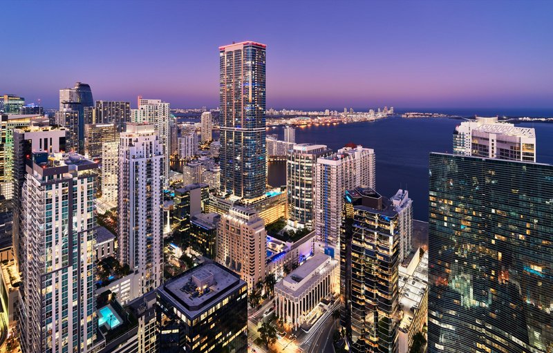 Aerial view of a modern high-rise city skyline at dusk with illuminated skyscrapers and a distant harbor