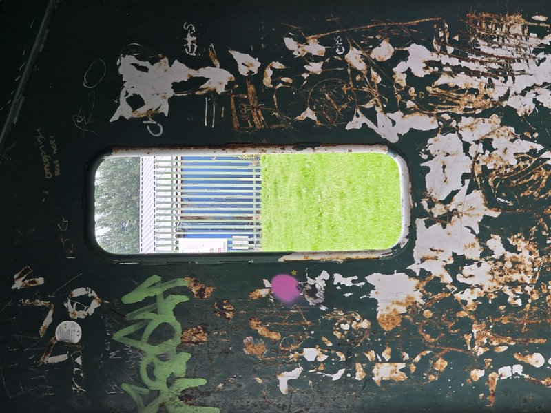Heavily rusted peeling dark green metal panel with graffiti frames a porthole window looking out to grass and fencing