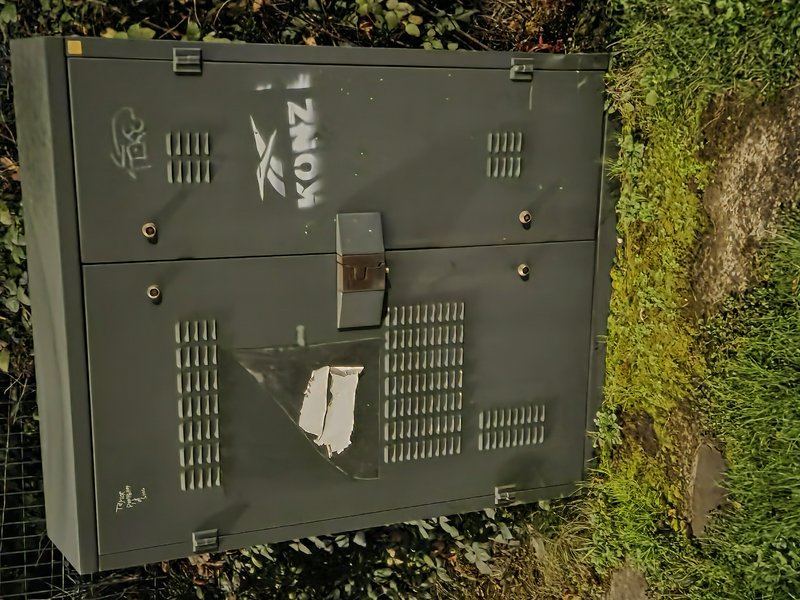 Dark grey vented outdoor electrical utility box covered in spray paint graffiti sits on mossy grassy ground