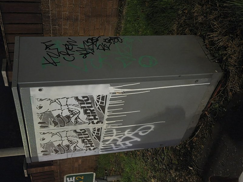 Gray outdoor utility box covered in overlapping spray paint graffiti and handwritten tag markings