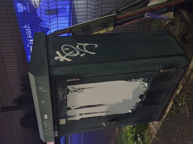 Tagged dark green electrical utility box sits next to a wire fence with a glowing blue car wash sign at night