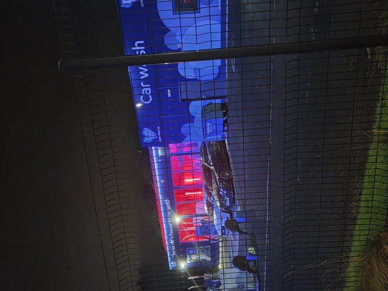 Illuminated automatic car wash at night with a sedan entering the bay, viewed through a wire security fence