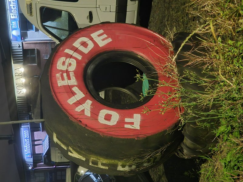 Oversized repurposed Fyreside tire marker rests on an overgrown roadside verge at night beside parked vehicles