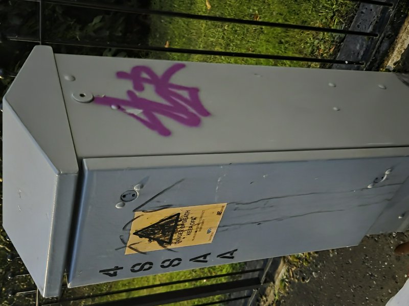 Grey street electrical utility box marked with purple graffiti and a defaced danger warning sticker