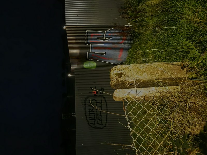 Night view of graffiti on a corrugated industrial building next to weathered fence posts and overgrown grass