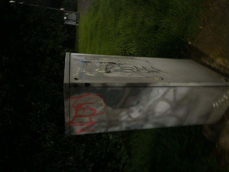 Motion-blurry dusk photo of a graffiti tagged metal utility box sitting on grass next to a street