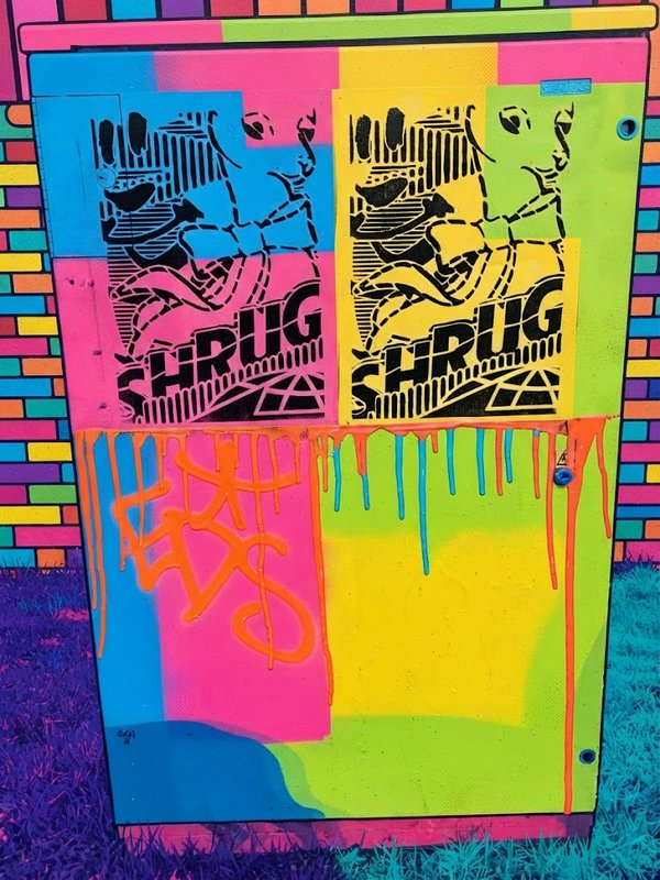 Bright neon painted utility box covered in street art shrug stencils and dripping paint details