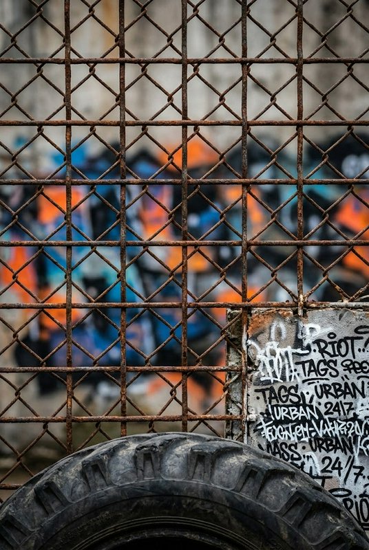 Rusted chain link fence sits in front of a graffiti covered wall with a heavy worn tire in the fo...