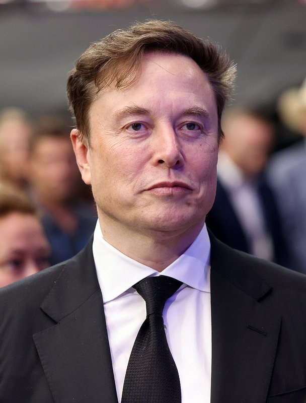 Close-up portrait of Elon Musk in a black suit and tie, with a neutral expression at a formal public event.