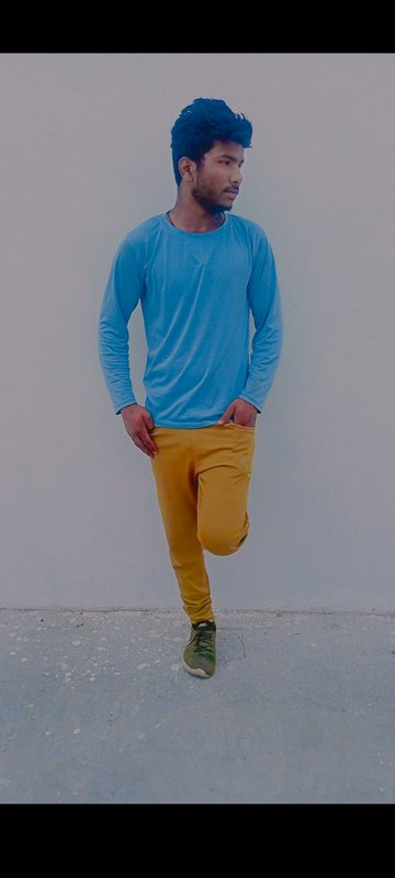 Young South Asian man poses casually against a plain wall, wearing a light blue long sleeve shirt and mustard yellow pants.