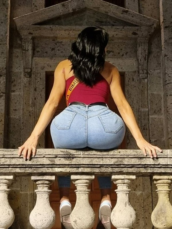 [Rear view of a woman with dark wavy hair sitting on an ornate weathered stone balcony, wearing a red halter top and skinn...
