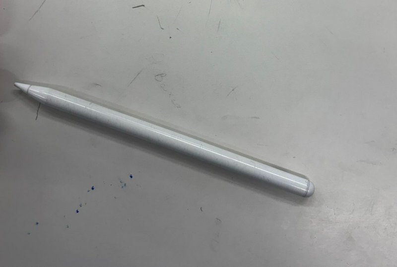 Glossy white Apple Pencil-style stylus resting diagonally on a scuffed light gray work desk surface