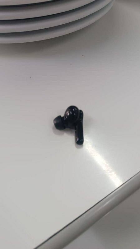 Single glossy black wireless TWS earbud resting on a reflective bright white table surface