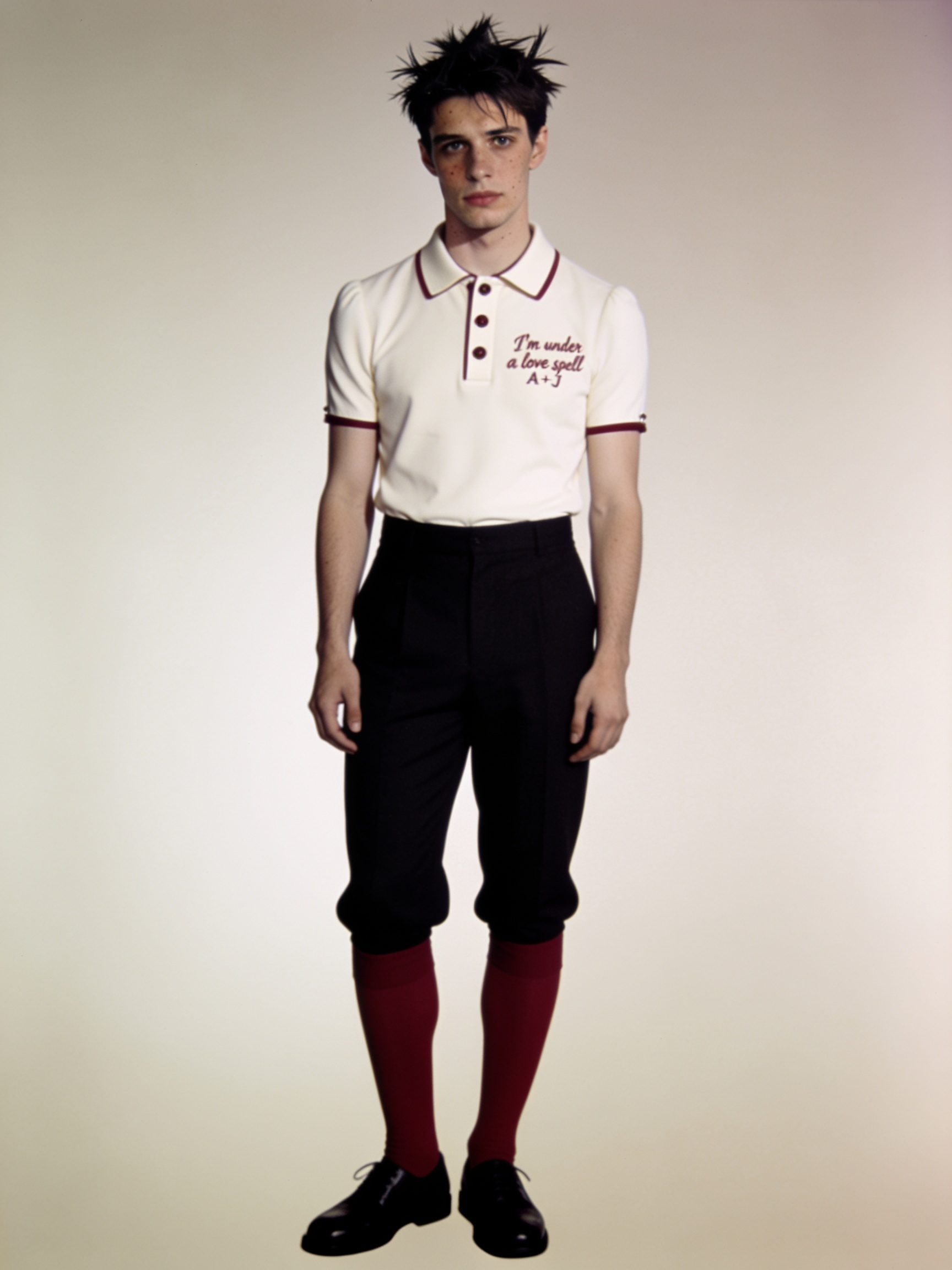 Young male model stands against a plain studio backdrop wearing a retro preppy outfit with embroidered cream ringer polo.