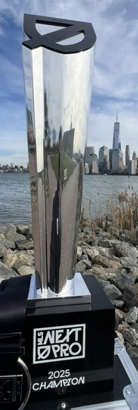 Reflective MLS Next Pro 2025 Champion trophy displayed against NYC Manhattan skyline waterfront b...