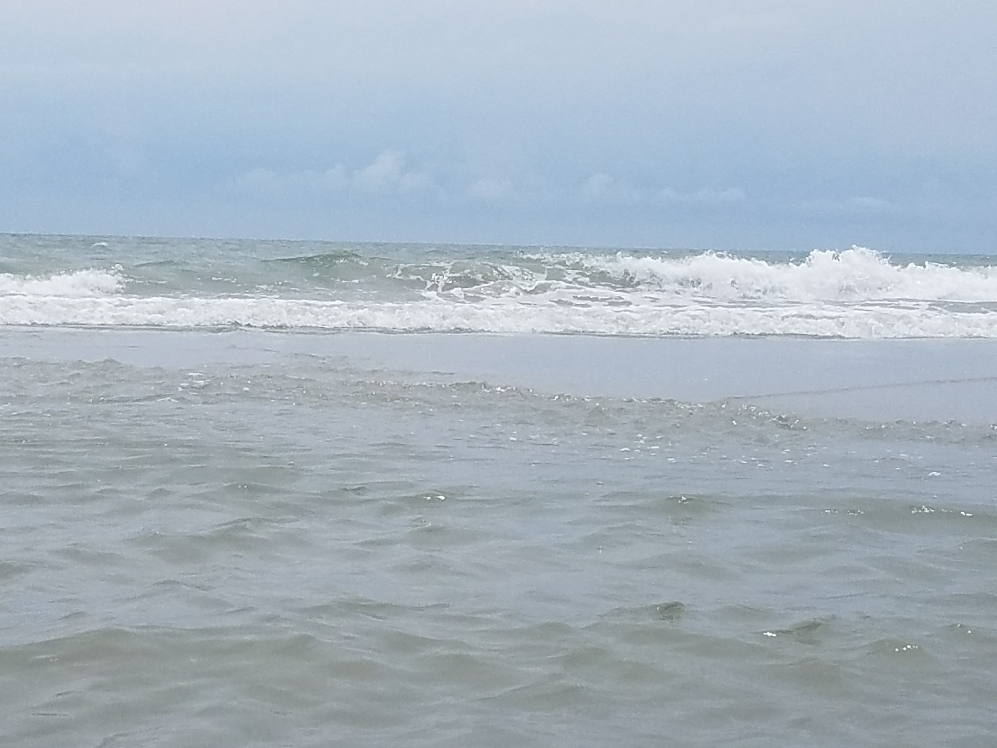 Rolling foamy ocean waves approach a shoreline under a pale overcast blue sky